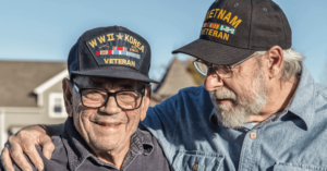 Two veterans stand next to one another outside and smile. Both are older men and wear black hats that say what wars they fought in.