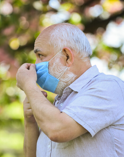 An older with white hair and a white beard stands outside and adjusts a blue face mask