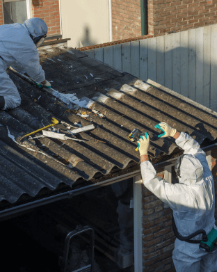 Two workers in white hazmat suits work to remove brown asbestos shingles from a roof
