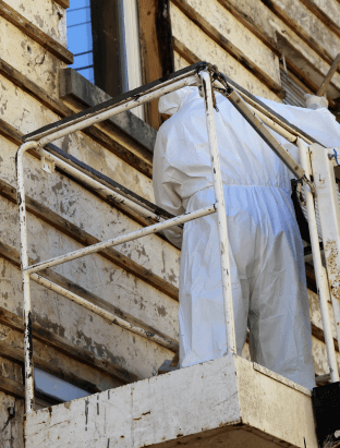 A worker in a white hazmat suit stands on a cream-colored scaffolding and works to remove asbestos from the side of a building