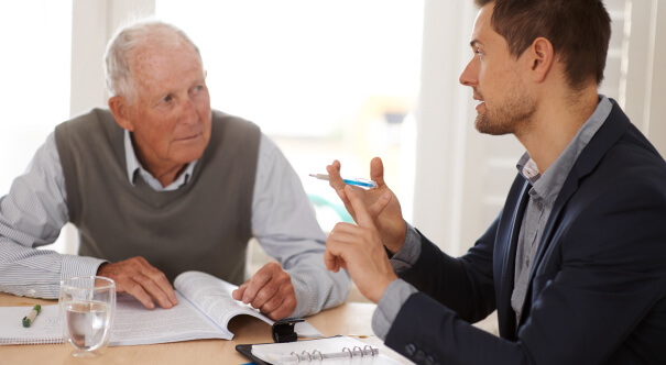An older man sits with a younger man at a table. Together, they review documents and engage in a discussion.