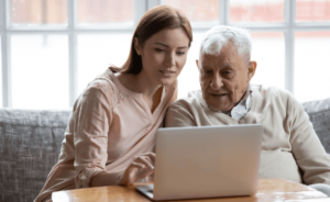 A young woman and an elderly male look at a laptop while sitting on a couch.