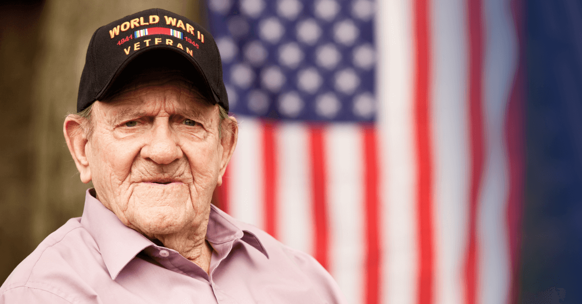 An older World War II veteran sits down. An American flag is in the background.