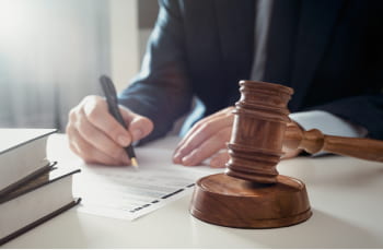 a man in a suit fills out a document on a desk. A gavel and books are also seen on the desk.