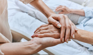 A younger person holds an older person's hand. The older person is shown to be sitting in bed.