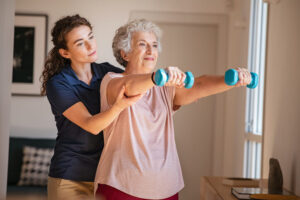 A younger woman serving as a physical therapist helps an older woman lift small weights.