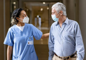 A nurse in scrubs walks with an older man in business casual attire. Both are wearing facemasks.