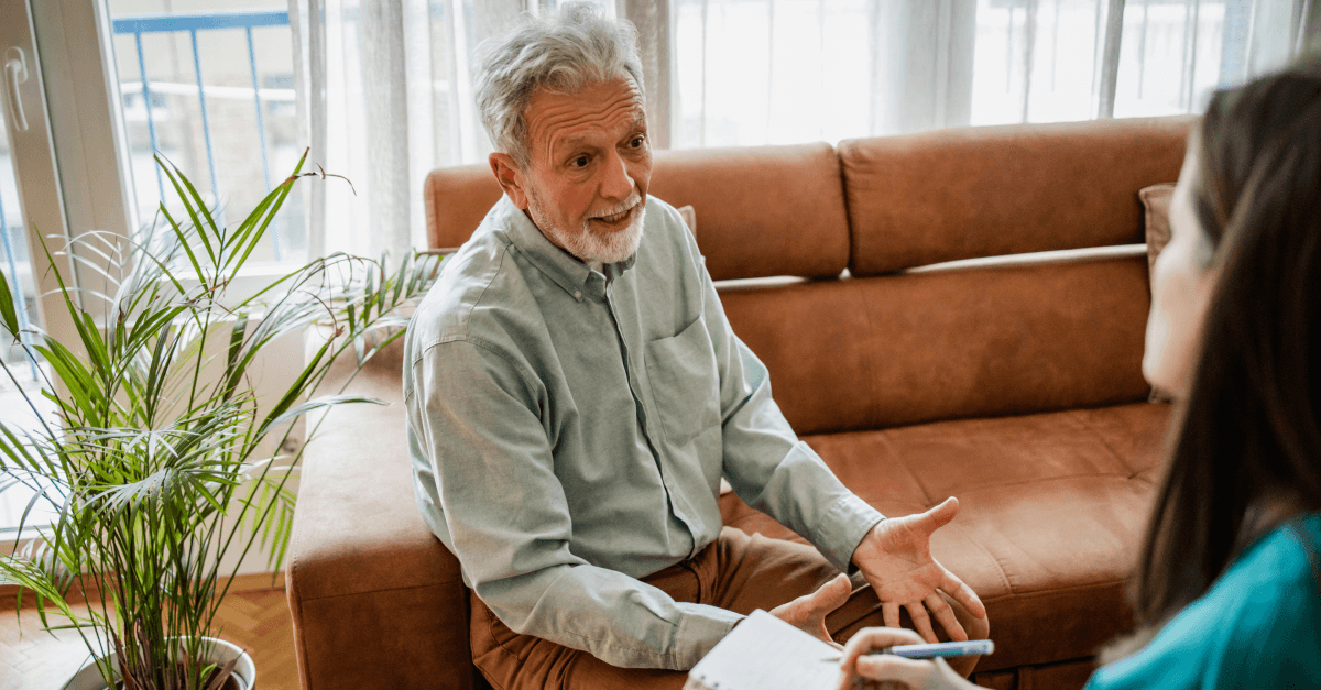 An older man sits on a couch while a younger woman approaches